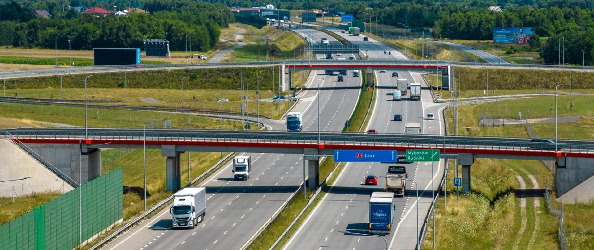 Autostrada A1 w okolicach Częstochowy z widocznymi wiaduktami i ruchem samochodów osobowych oraz ciężarowych, otoczona terenami zielonymi i zabudową w oddali.