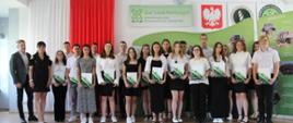 Grupa uczniów wraz z dwójką dorosłych stoi w dwóch rzędach uczniowie trzymają w ręku świadectwa szkolne w biało zielonych teczkach