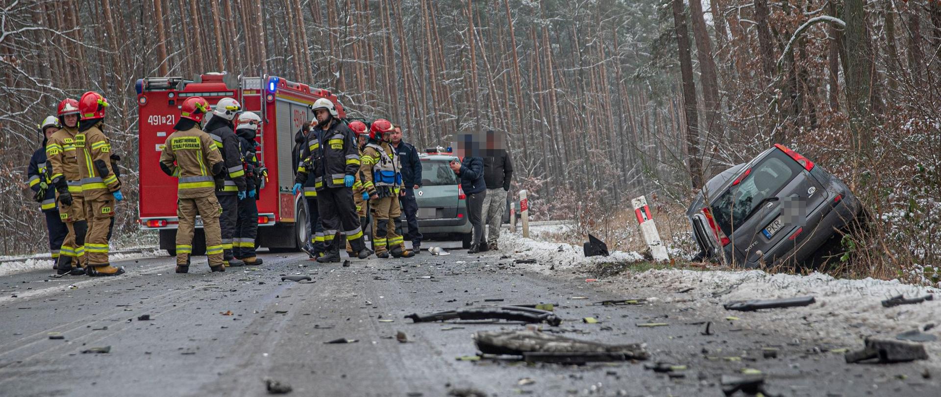 Zdjęcie przedstawia samochód strażacki koloru czerwonego, stojących strażaków i policjantów przy rozbitym samochodzie leżącym w rowie podczas wykonywania swoich czynności.