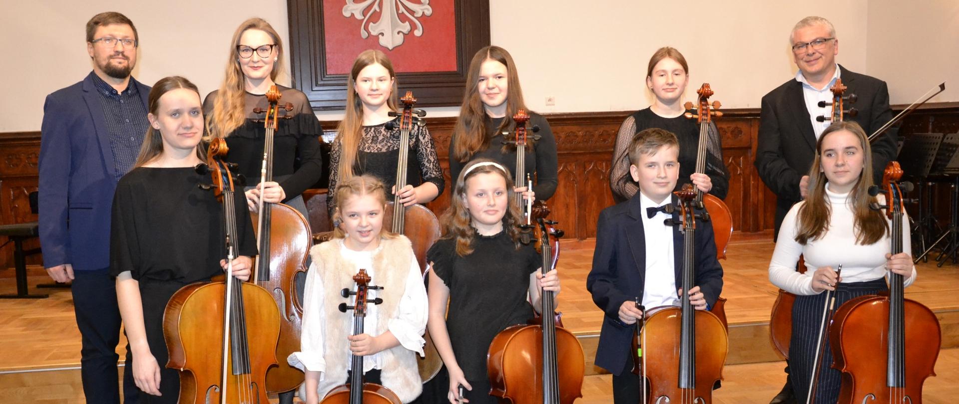 zdjęcie grupowe przedstawia uczniów klas wiolonczeli naszej szkoły trzymających swoje instrumenty, stojąc na scenie sali królewskiej