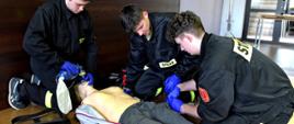 Zdjęcie zrobione w sali szkoleniowej podczas kursu kwalifikowanej pierwszej pomocy. Trzech strażaków ćwiczy udzielanie pierwszej pomocy na fantomie podłączonym defibrylatora. 