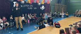 
Jeden ze strażaków trzyma mikrofon i przemawia do siedzących dzieci. Strażacy prezentują udzielanie pierwszej pomocy.
