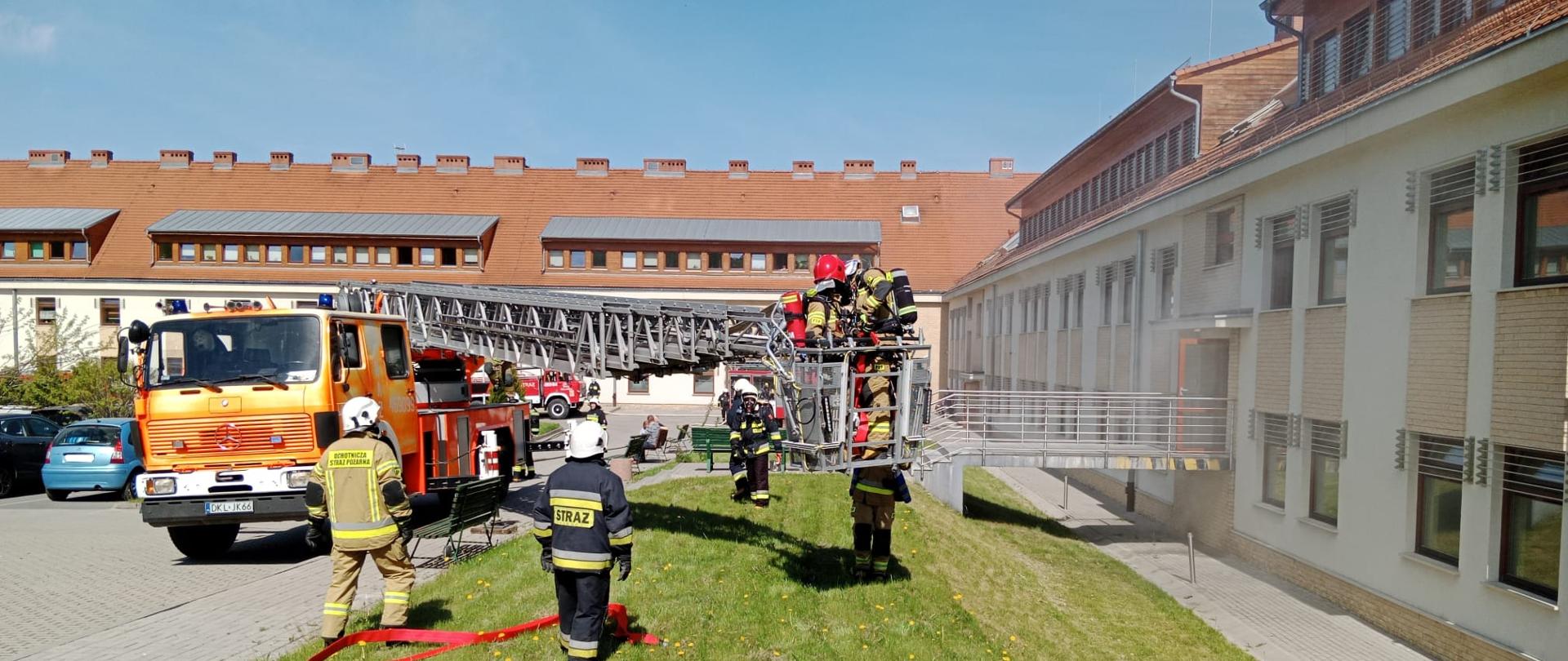 Plac przed budynkiem szpitala. W koszu podnośnika stoi dwóch strażaków. Wokół podnośnika stoją strażacy biorący udział w ćwiczeniach.