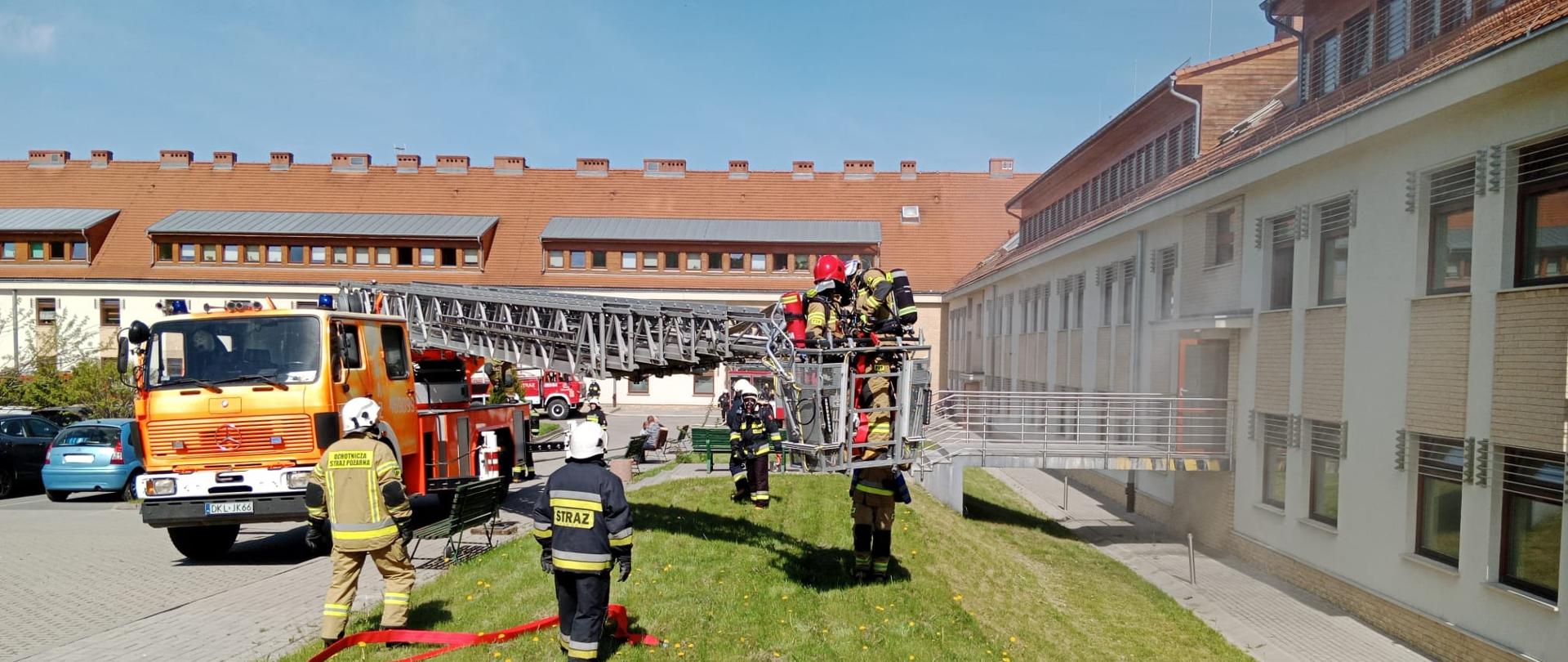Plac przed budynkiem szpitala. W koszu podnośnika stoi dwóch strażaków. Wokół podnośnika stoją strażacy biorący udział w ćwiczeniach.