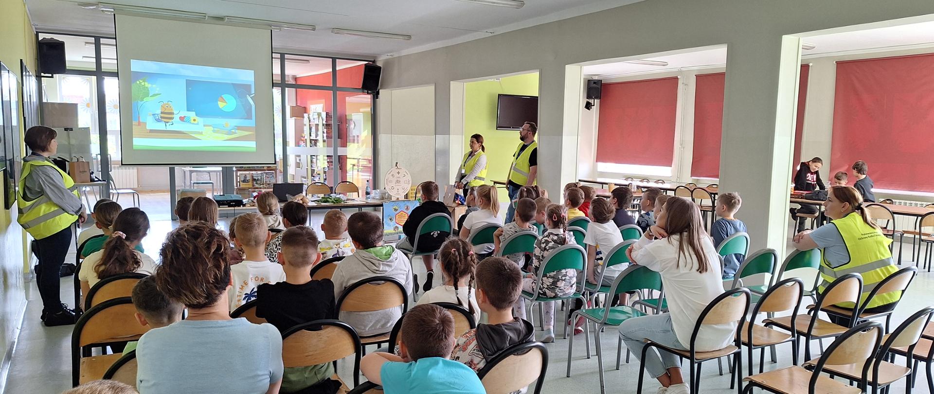 Zdjęcie przedstawia salę wypełnioną dziećmi. Dzieci siedzą na krzesłach i oglądają prezentację wyświetlaną na ekranie do prezentacji.