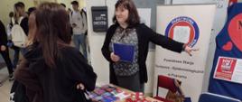 Przy stoisku z materiałami edukacyjnymi i upominkami walentynkowymi stoi kobieta oraz grupa uczniów. Kobieta wskazuje tablicę.