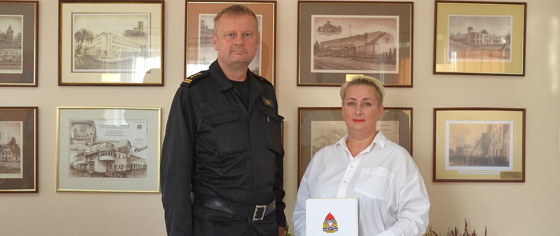 Zdjęcie przedstawia komendanta powiatowego PSP i Arletę Rueck z listem gratulacyjnym. W tle grafiki o tematyce PSP