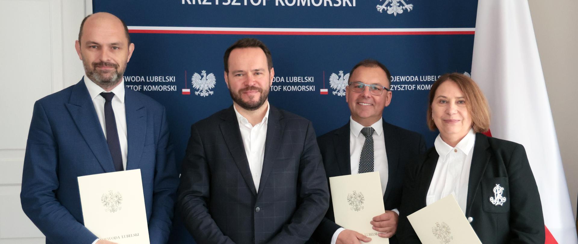 Cztery osoby w eleganckich strojach stoją w rzędzie, trzymając w rękach oficjalnie wyglądające dokumenty z pieczęciami. Za nimi widnieje biało-czerwony baner z tekstem "WOJEWODA LUBELSKI" i "KRZYSZTOF KOMORSKI" nad godłem Polski - białym orłem w koronie na czerwonym tle. Po prawej stronie widoczny jest fragment polskiej flagi. Osoby te wydają się pozować do zdjęcia po oficjalnym wydarzeniu lub ceremonii.