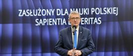 Wiceminister Murdzek stoi i mówi do mikrofonu na stojaku, za nim wbudowany w ścianę wielki ekran z napisem Ceremonia wręczenia medali „Zasłużony dla Nauki Polskiej Sapientia et Veritas”.