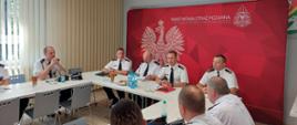 Zdjęcie przedstawia przemawiającego Zastępcę Wielkopolskiego Komendanta Wojewódzkiego. Obok siedzą Komendant Miejski PSP wraz z zastępcą oraz Prezes Zarządu Oddziału Powiatowego ZOSP RP. W tle ścianka komendy z godłem.
