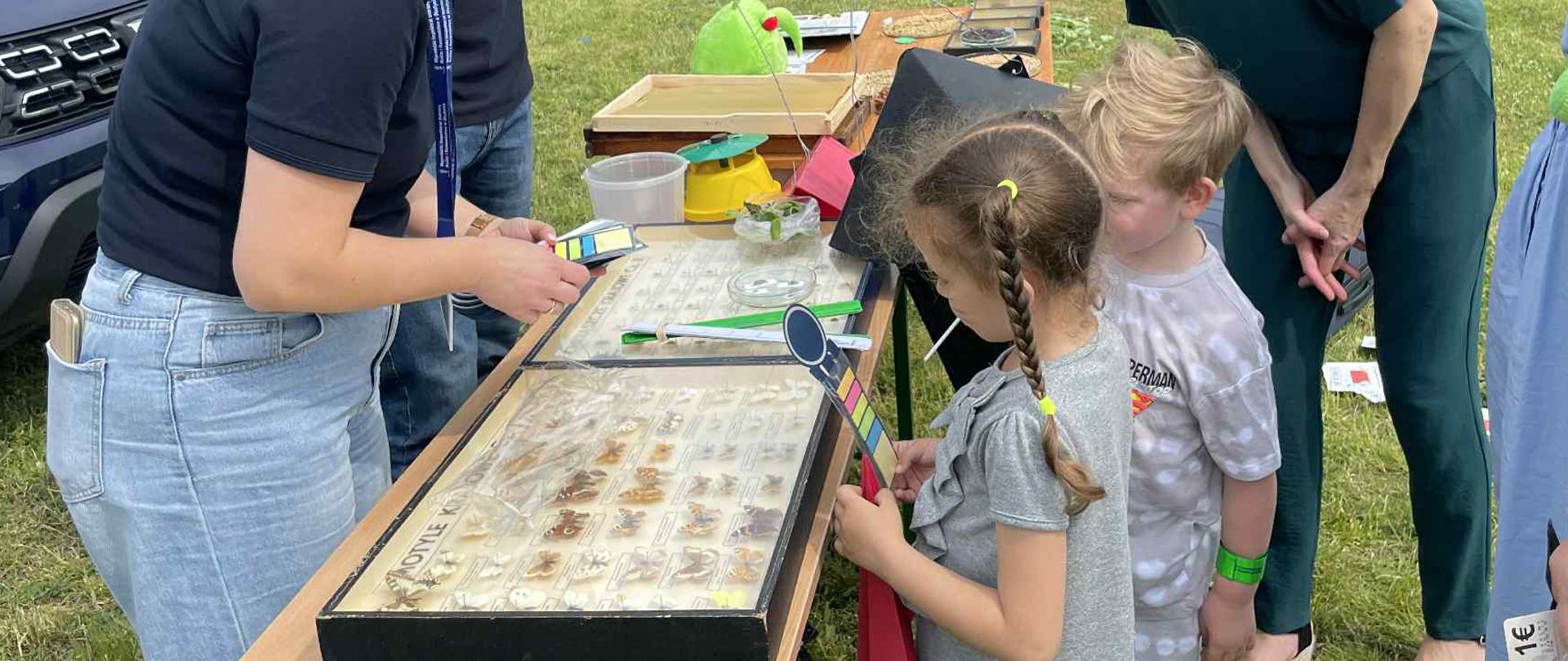 stolik edukacyjny ustawiony na trawie, przy którym prowadzone są zajęcia dla dzieci. Dwoje dorosłych prowadzących, ubranych w granatowe koszulki, pokazuje dzieciom eksponaty. Dzieci z zainteresowaniem przyglądają się materiałom, towarzyszą im opiekunowie