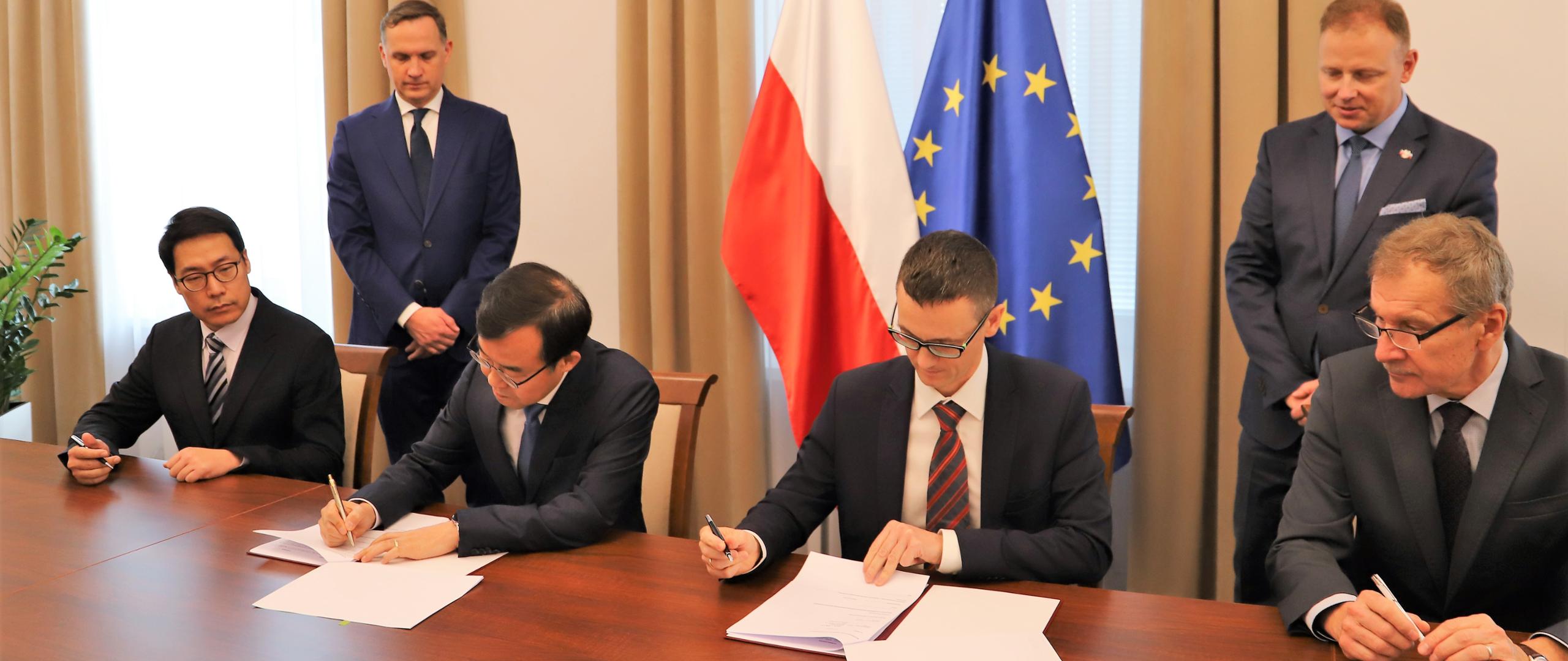 Dwóch mężczyzn siedzących przy stole i podpisujących porozumienie. W tle flagi Polski i UE.