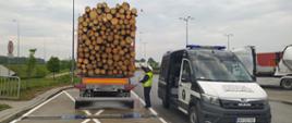 Ciężarówka przewożąca drewno stoi tuż za przenośnymi wagami. Inspektor mierzy wysokość naczepy z ładunkiem. Po prawej stronie stoi oznakowany radiowóz ITD typu furgon.