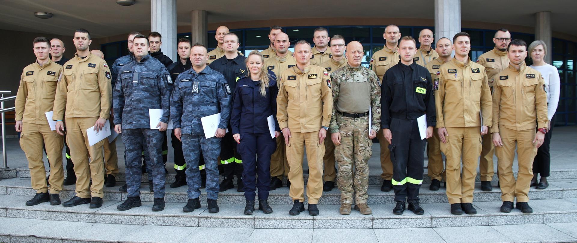 Pamiątkowe zdjęcie grupowe funkcjonariuszy PSP, Policji oraz Straży Marszałkowskiej (21 osób) w towarzystwie Zastępcy Komendanta Centralnej Szkoły PSP, kobiety oraz trzech funkcjonariuszy CS PSP