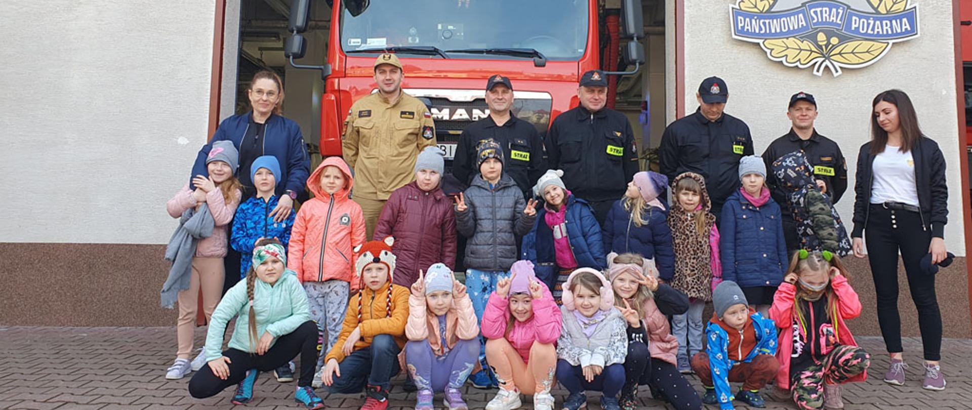 Na zdjęciu widać dzieci ze szkoły podstawowej w obecności zastępcy komendanta powiatowego, nauczycielek oraz zmiany służbowej na tle samochodu gaśniczego i garaży jrg.