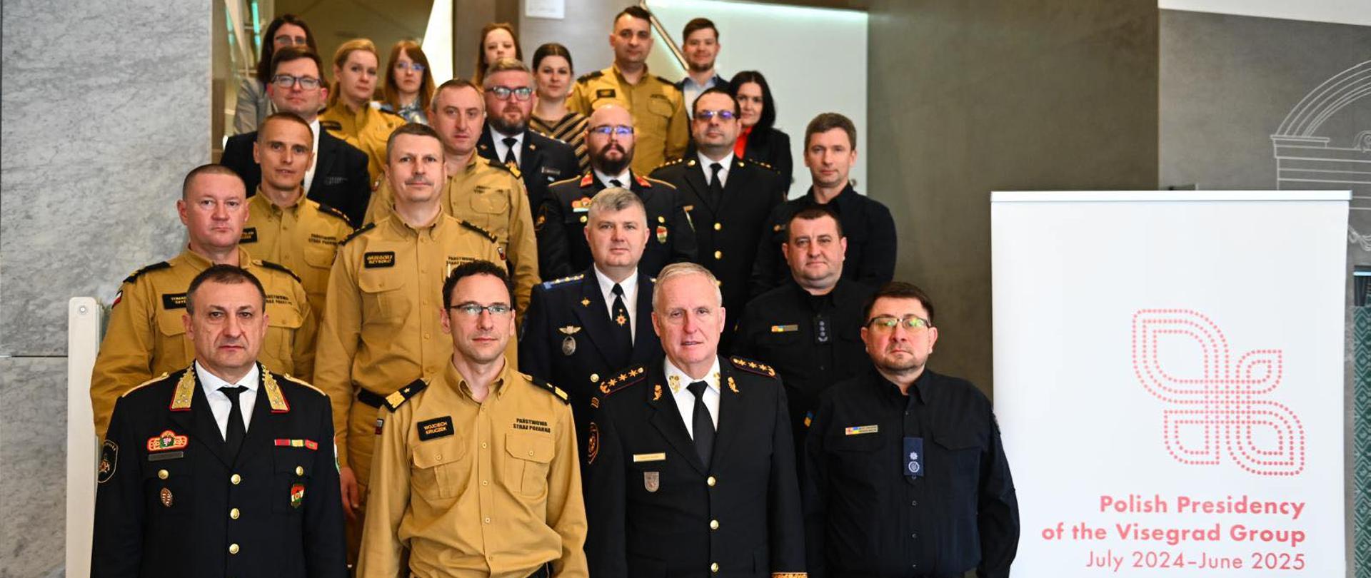 Zdjęcie grupowe wszystkich uczestników konferencji, w tym funkcjonariuszy PSP w żółtych mundurach służbowych, zagranicznych przedstawicieli służb ratowniczych oraz osób cywilnych, wykonane na schodach budynku obrad, obok banera z napisem „Polish Presidency of the Visegrad Group”.
