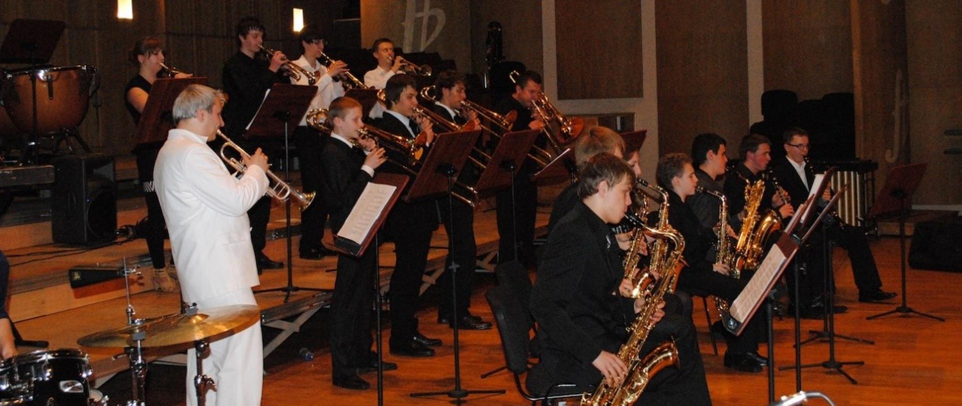 Grupa uczniów grająca na instrumentach dętych. W pierwszy rzędzie siedzą przy pulpitach saksofoniści. W drugim i trzecim stoją trębacze i puzoniści. Wszyscy ubrani są w ciemne garnitury lub białe koszule. Obok stoi pedagog w białym garniturze grający na trąbce.