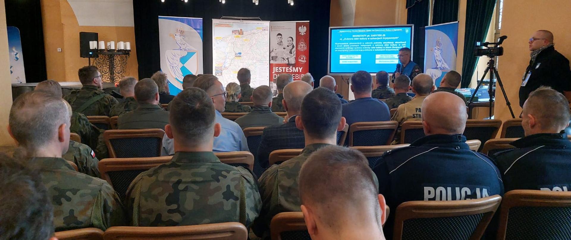 Duża sala konferencyjna, wypełniona uczestnikami w mundurach wojskowych i policyjnych. Na scenie za stołem prezydialnym stoją banery z orłem, mapą oraz plakatem z napisem 'Jesteśmy dla Ciebie'. Uczestnicy odwróceni tyłem, skierowani w stronę prelegenta prezentującego informacje na ekranie