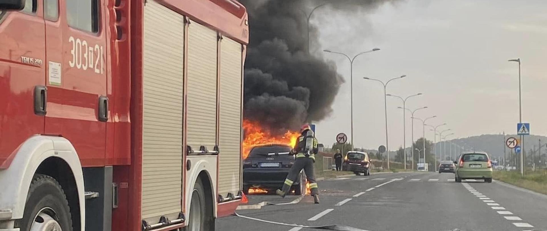 Zdjęcie przedstawia ulicę Krakowską. Po lewej stoi samochód pożarniczy zza którego widać palący się samochód osobowy. Przed pojazdem przechodzi strażak z linia i prądownicą pianową. W głębi widać przypadkowe auta.
