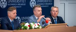 Minister Wieczorek siedzi za stołem, mówi do kilku mikrofonów, obok niego siedzi dwóch mężczyzn w garniturach, za nimi granatowa ścianka z napisem Uniwersytet Kazimierza Wielkiego w Bydgoszczy.