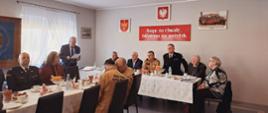 Strażacy Państwowej Straży Pożarnej oraz osoby cywilne siedzą za stołami na których rozłożony jest poczęstunek.