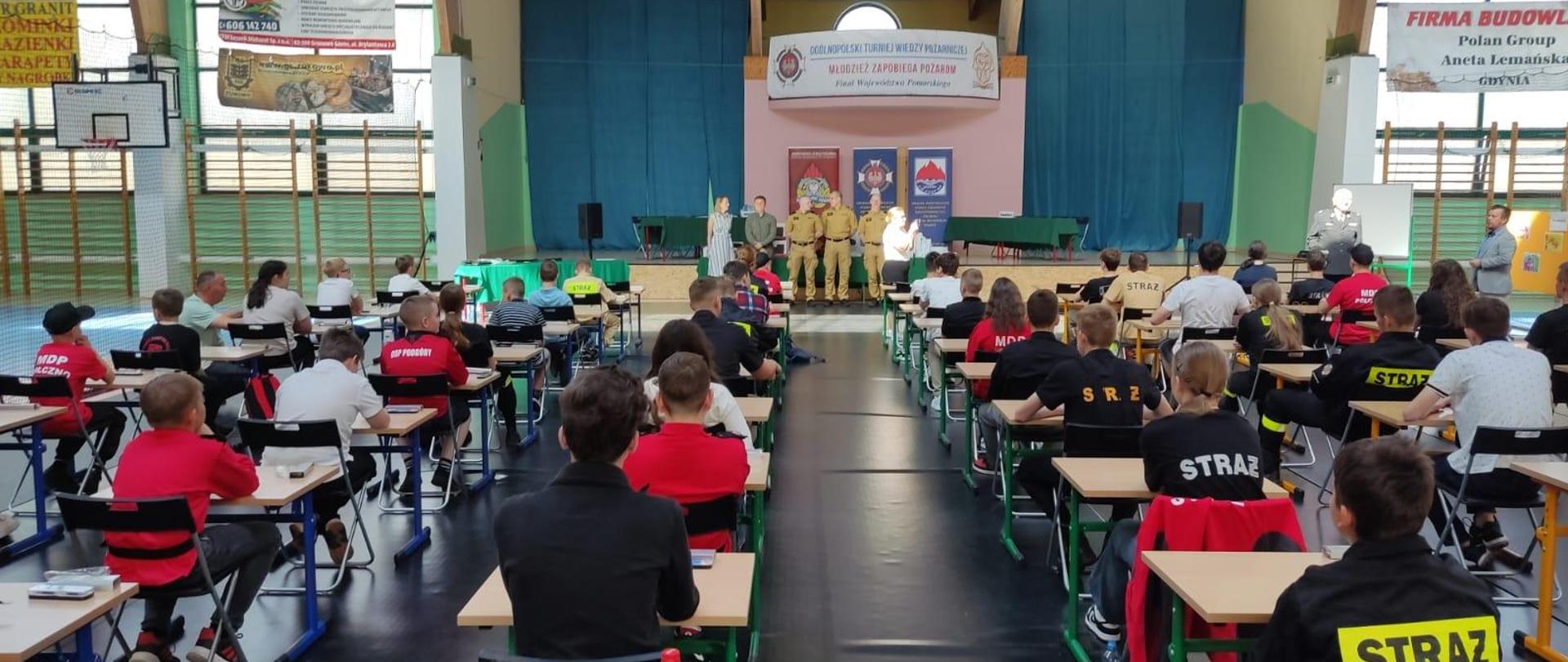 Zdjęcie przedstawia uczestników konkursu pożarniczego siedzących w ławkach.