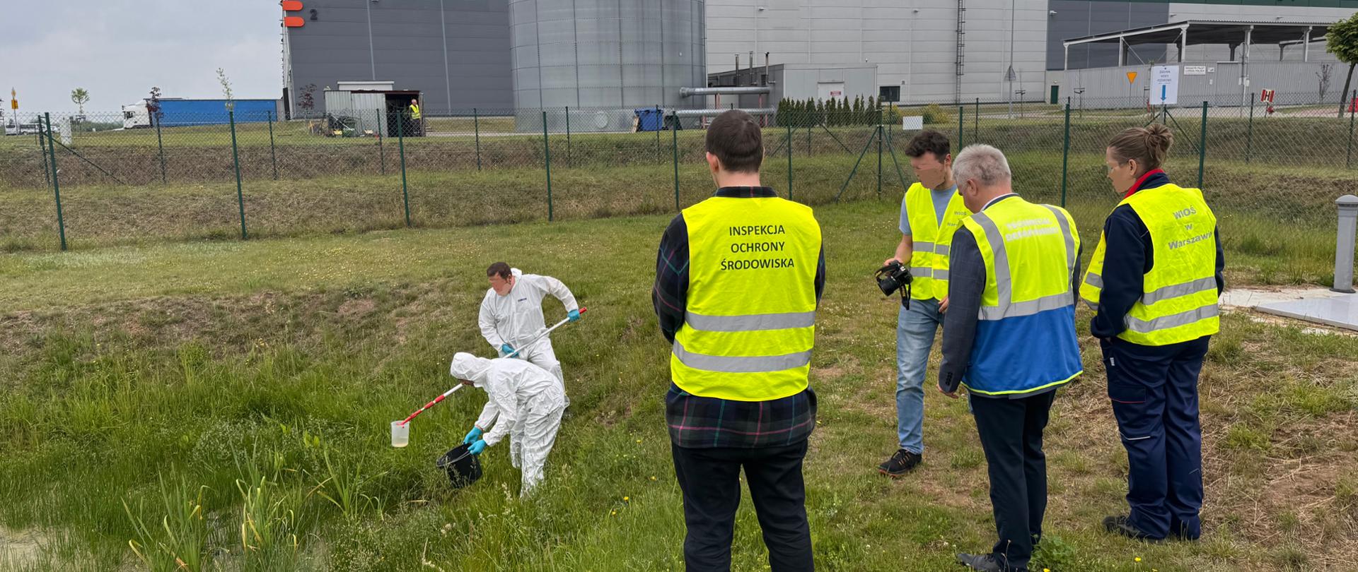 Na otwartym, porośniętym trawą terenie, stoi troje inspektorów Wojewódzkiego Inspektoratu Ochrony Środowiska w Warszawie oraz przedstawiciel zakładu przemysłowego. Monitorują oni pobór próbek dokonywany przez dwóch pracowników Centralnego Laboratorium Badawczego GIOŚ ubranych w białe ochronne stroje. W tle widać szarą halę.