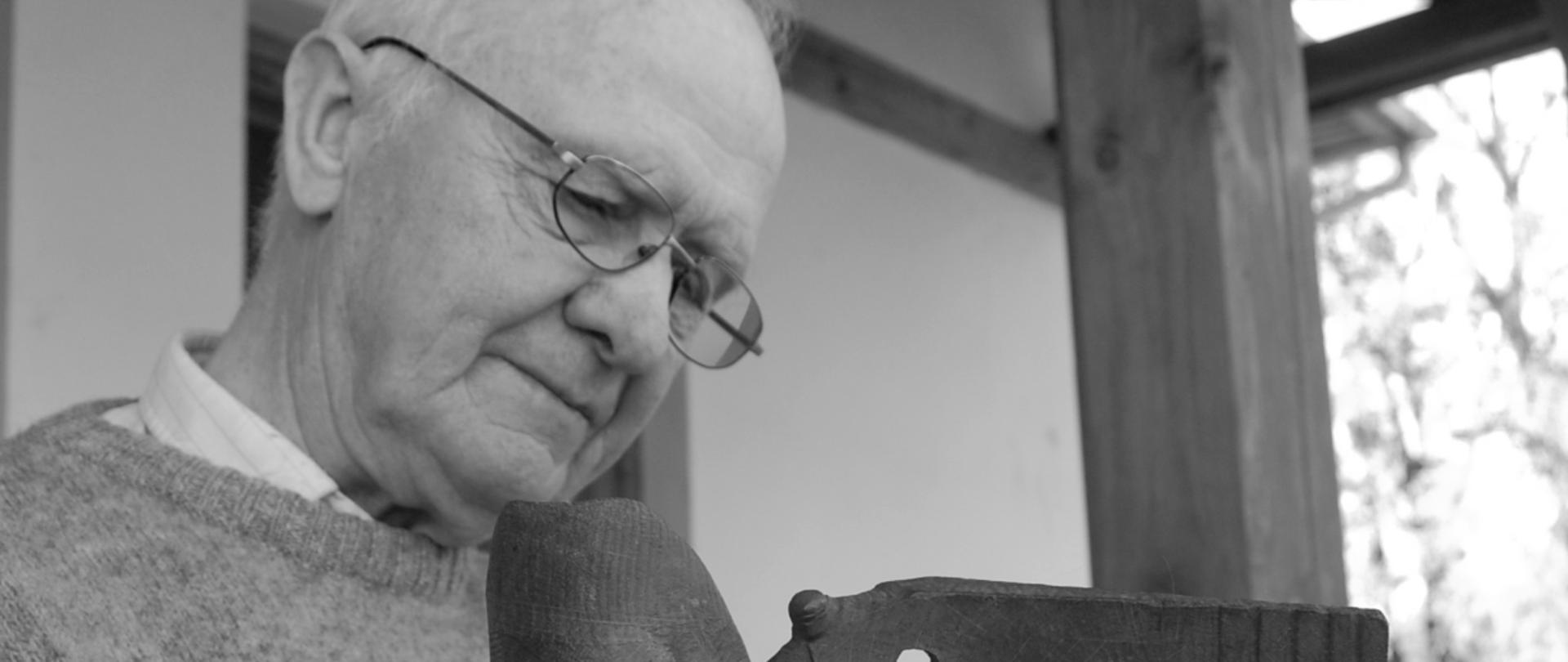 Na zdjęciu znajduje się postać mężczyzny. Jest to Antoni Grabowski - Artysta Rzeźbiarz wieloletni pedagog Państwowego Liceum Sztuk Plastycznych im. Antoniego Kenara w Zakopanem. Artysta jest ubrany w szary sweter, ma założone okulary. W prawej ręce trzyma własną rzeźbę wykonaną z drewna. W tle znajduje się fragment budynku szkoły - warsztatów przy ul. Kościeliskiej 35 w Zakopanem.