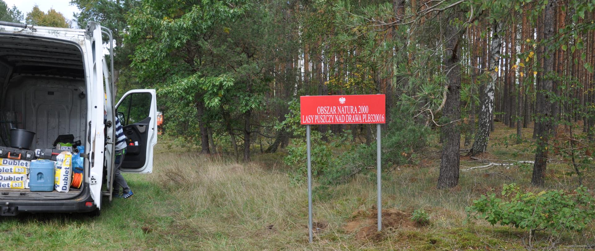Po lewej stronie fragment białego samochodu z otwartym bagażnikiem. Po prawej stronie czerwona tablica z napisem Obszar Natura 2000 Lasy Puszczy nad Drawą. W tle fragment lasu.