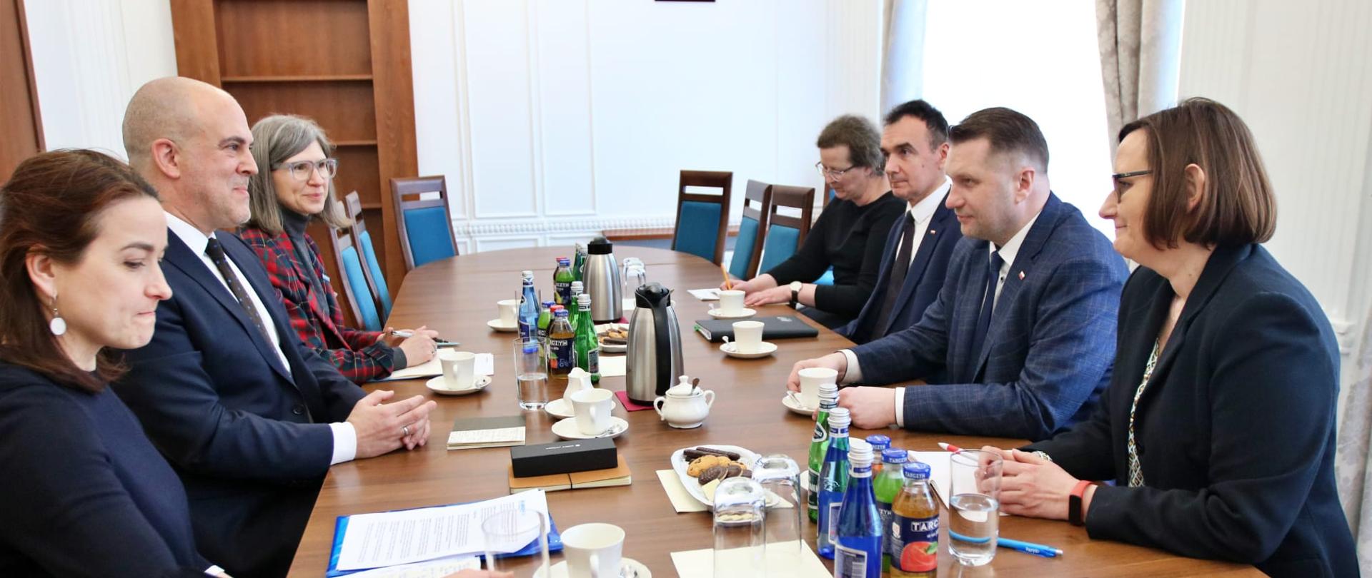 Zdjęcie z boku, po dwóch stronach podłużnego drewnianego stołu siedzi siedem rozmawiających osób, za stołem biała ściana i drewniana szafka z pustymi półkami, z prawej zasłonięte firankami okno.