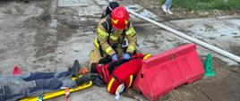 Strażak przy torbie medycznej, obok poszkodowana osoba ułożona na ziemi, w tle samochody gaśnicze i pracownicy firmy.