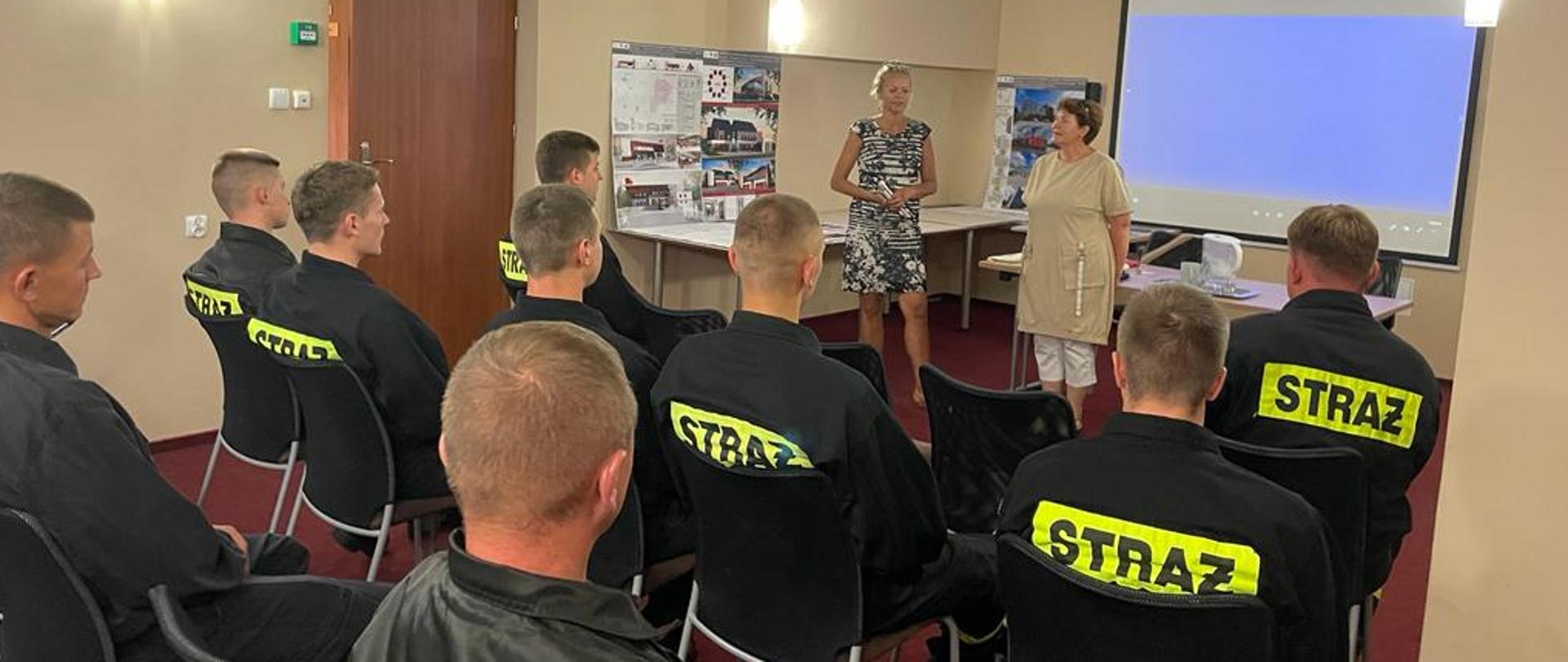 W pomieszczeniu prze dwiema kobietami siedzi tyłem kilku strażaków w czarnych mundurach.