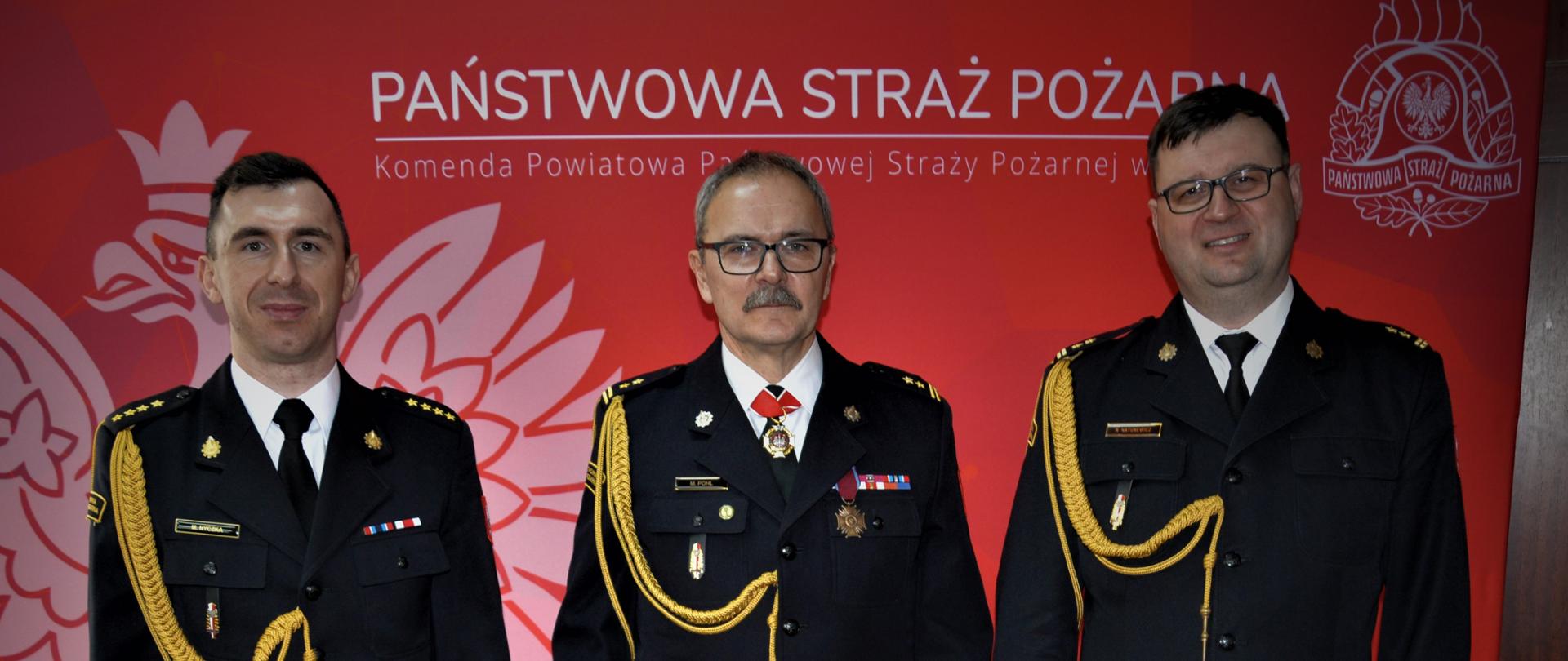 Zdjęcie przedstawia 3 osoby pozujące do zdjęcia. Są to: Zastępca Wielkopolskiego Wojewódzkiego Komendanta Wojewódzkiego PSP w Poznaniu, Komendant Powiatowy PSP w Gostyniu oraz Zastępca Komendanta Powiatowego PSP w Gostyniu. Ubrani są w umundurowanie wyjściowe.
