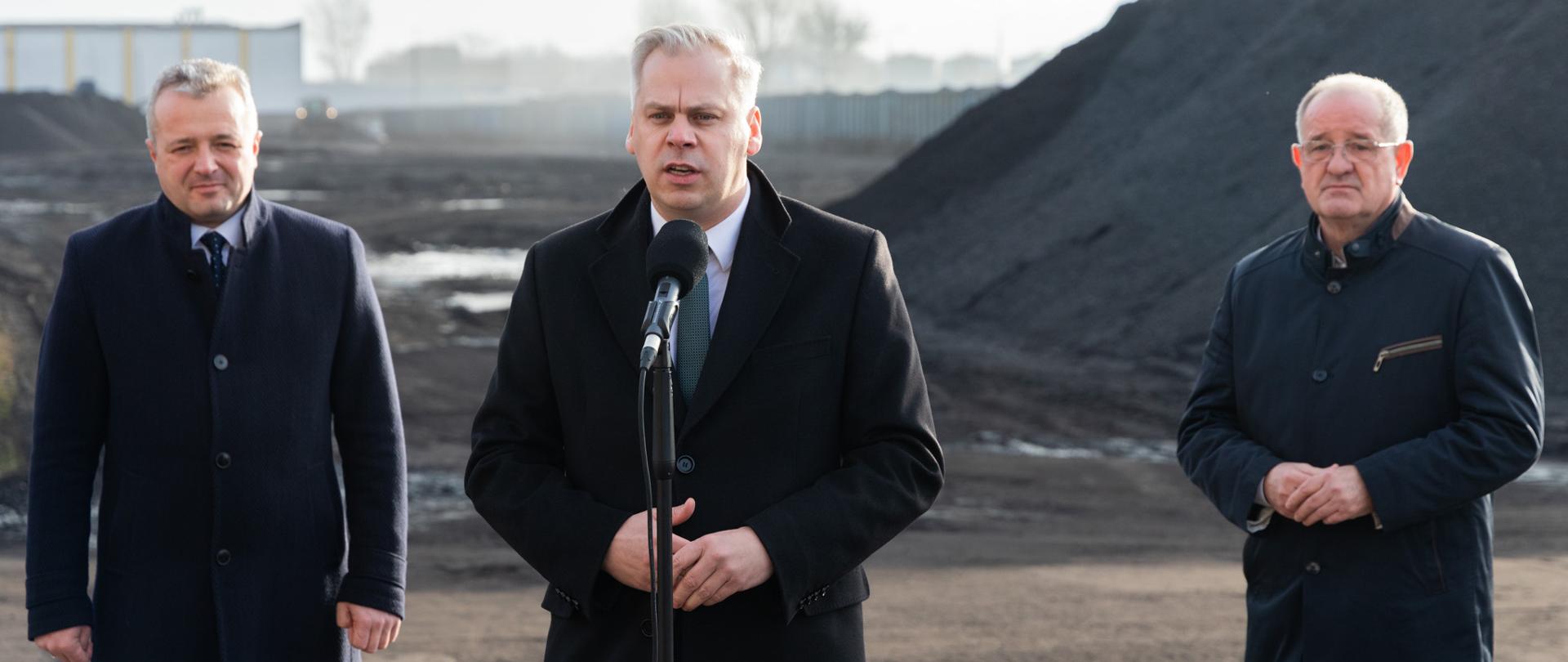 Wiceminister Karol Rabenda stoi za mikrofonem. Z tyłu stoi wojewoda kujawsko-pomorski i prezes firmy TOREC Waldemar Kozieł. W tle hałdy węgla. 