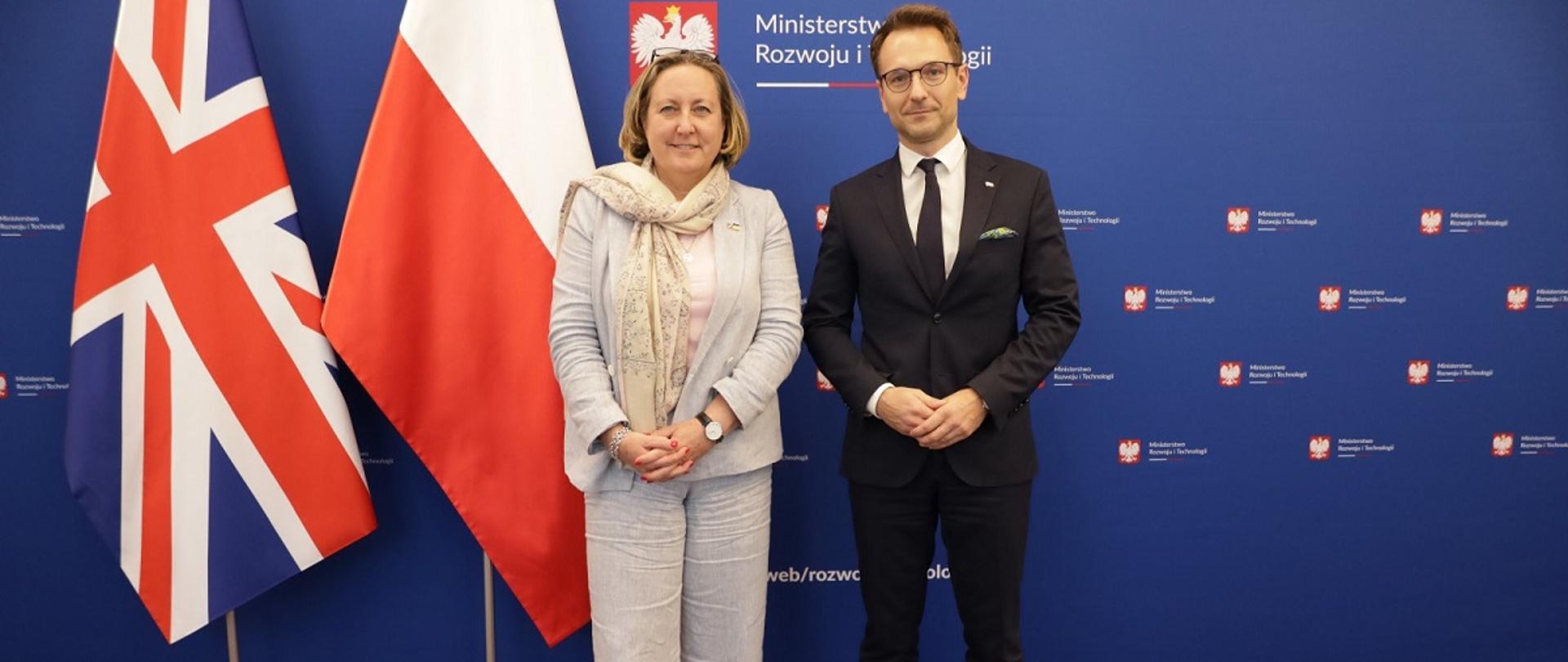 Minister rozwoju i technologii Waldemar Buda i minister handlu międzynarodowego Wielkiej Brytanii Anne-Marie Trevelyan, ministrowie stoją na tle flag Polski i Wielkiej Brytanii, w tle banner z logo MRiT