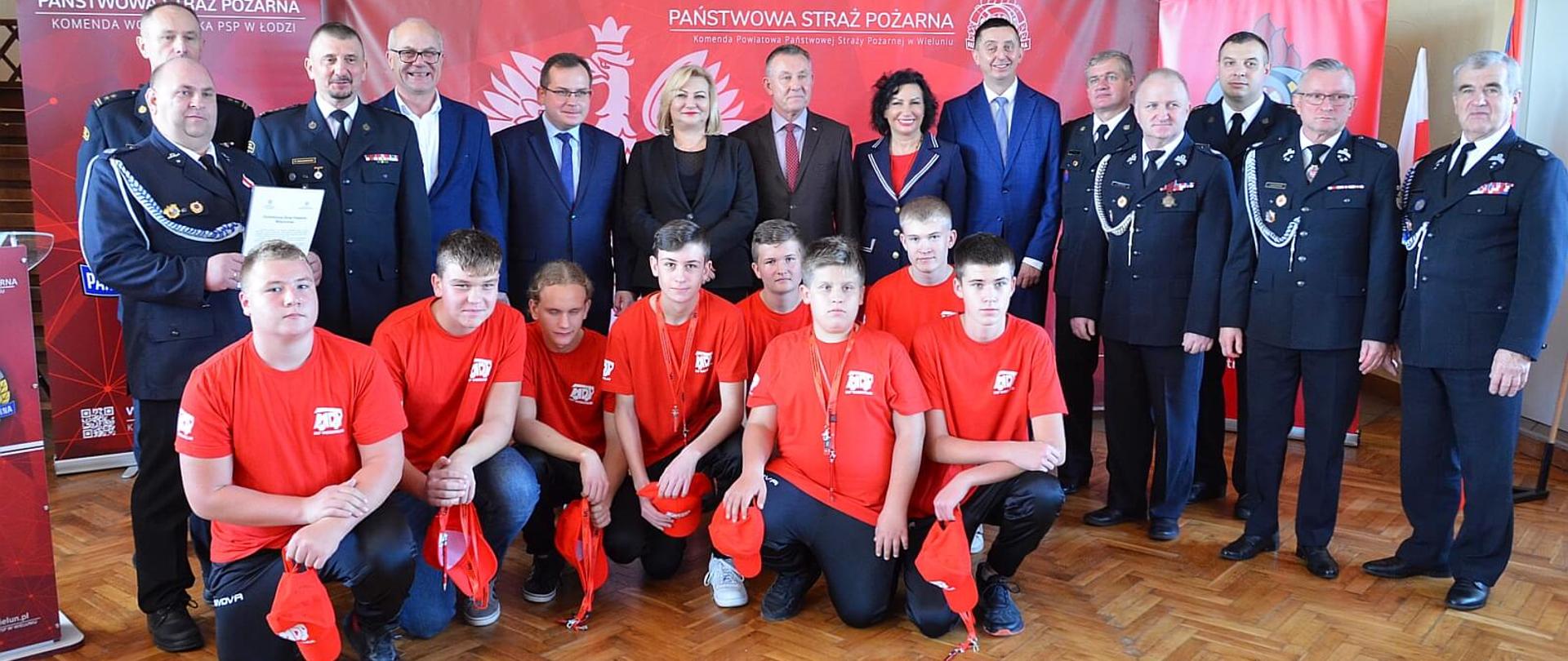 uczestnicy uroczystości wręczenia promes dla młodzieżowych drużyn pożarniczych pozują do zdjęcia zbiorowego wśród nich dzieci z młodzieżowej drużyny pożarniczej, strażacy druhowie, posłowie i senatorowie RP, przedstawiciele władz samorządowych