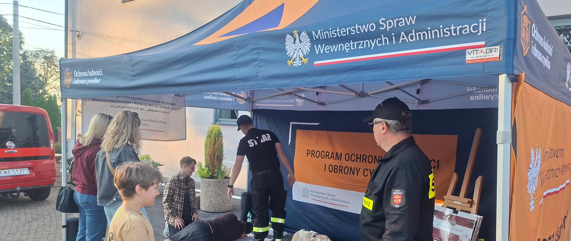 Na zdjęciu widać namiot Akademii OLiOC ze stanowiskami edukacyjnymi z zakresu poradnika bezpieczeństwa plecaka ewakuacyjnego i pierwszej pomocy oraz strażaka prowadzącego szkolenie dla ludności. 