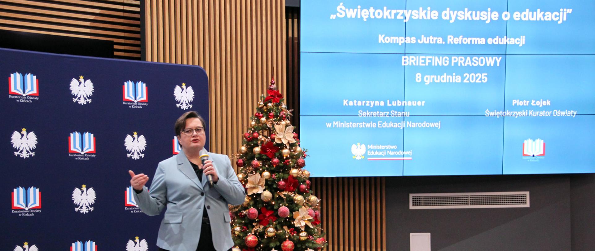 Wiceminister Katarzyna Lubnauer, ubrana w błękitną marynarkę i czarne spodnie, przemawia przez mikrofon ze sceny do uczestników wydarzenia. W tle widać ściankę Kuratorium Oświaty w Kielcach, choinkę świąteczną oraz ekran z napisem "Świętokrzyskie dyskusje o edukacji", datą wydarzenia oraz gośćmi konferencji.