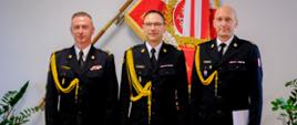 Trzech mężczyzn - strażaków w ciemnych mundurach wyjściowych stoi w rzędzie na tle flagi Polski i innej czerwono-białej flagi z symbolem w gabinecie komendanta głównego Państwowej Straży Pożarnej. Od lewej stoi st. bryg. Damian legierski śląski komendant wojewódzki Państwowej Straży Pożarnej. Jego mundur jest ozdobiony złotym sznurem. W środku stoi nadbryg. Wojciech Kruczek komendant główny Państwowej Straży Pożarnej. Po prawej stronie stoi mł. bryg. Zbigniew Dyk nowo powołany zastępca śląskiego komendanta wojewódzkiego Państwowej Straży Pożarnej, w ręku trzyma biały dokument -akt powołania na stanowisko Wszyscy trzej mężczyźni mają na sobie białe koszule i ciemne krawaty. Za nimi widać fragment rośliny doniczkowej po lewej i prawej stronie.