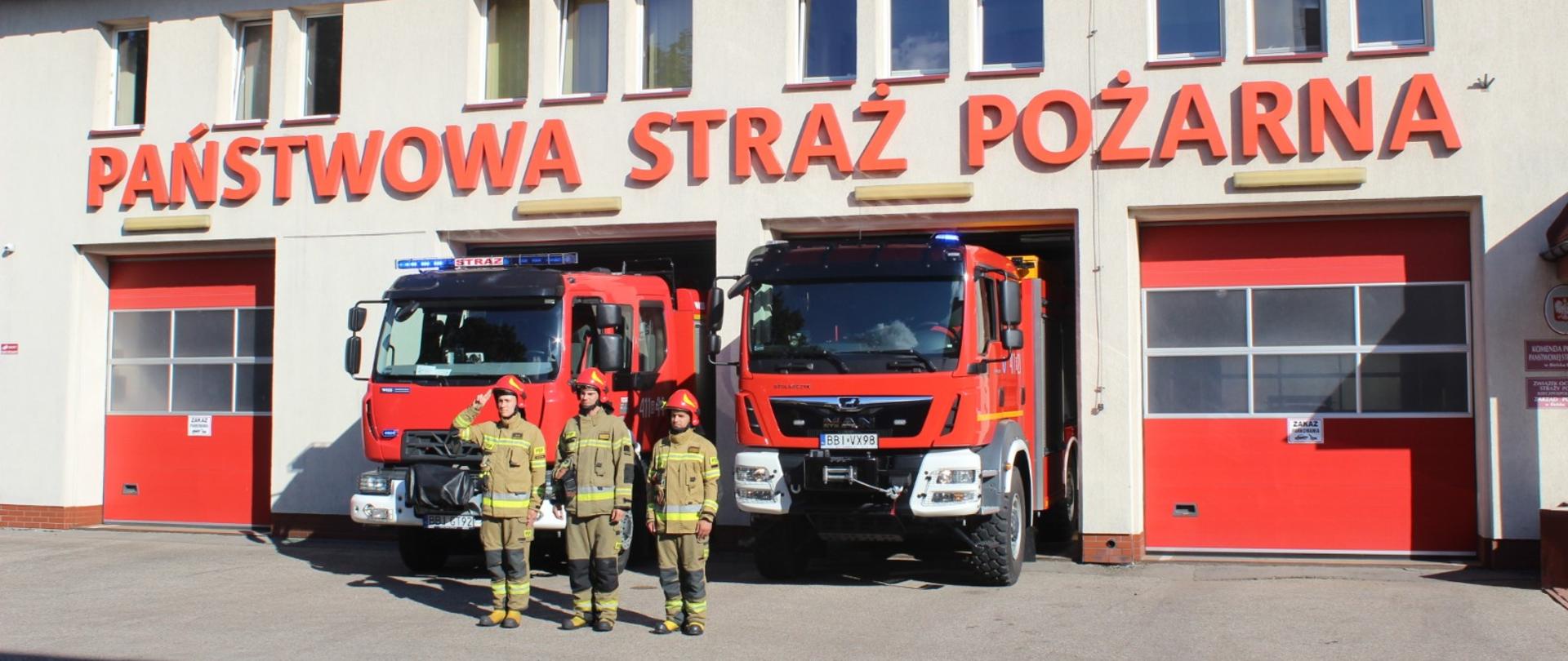 Zdjęcie przedstawia strażaków w ubraniach specjalnych przed budynkiem Jednostki Ratowniczo-Gaśniczej 