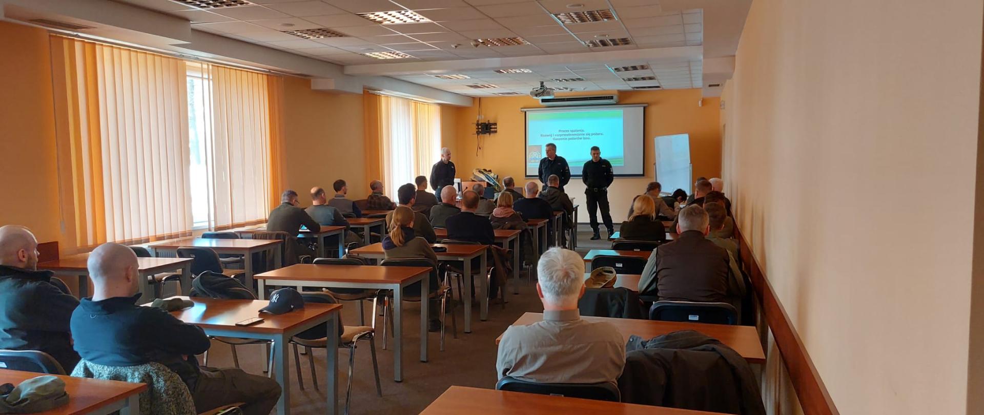 Uczestnicy szkolenia siedzą w ławkach. W tle widać prowadzących, którzy stoją przy tablicy, na której wyświetlana jest prezentacja,
