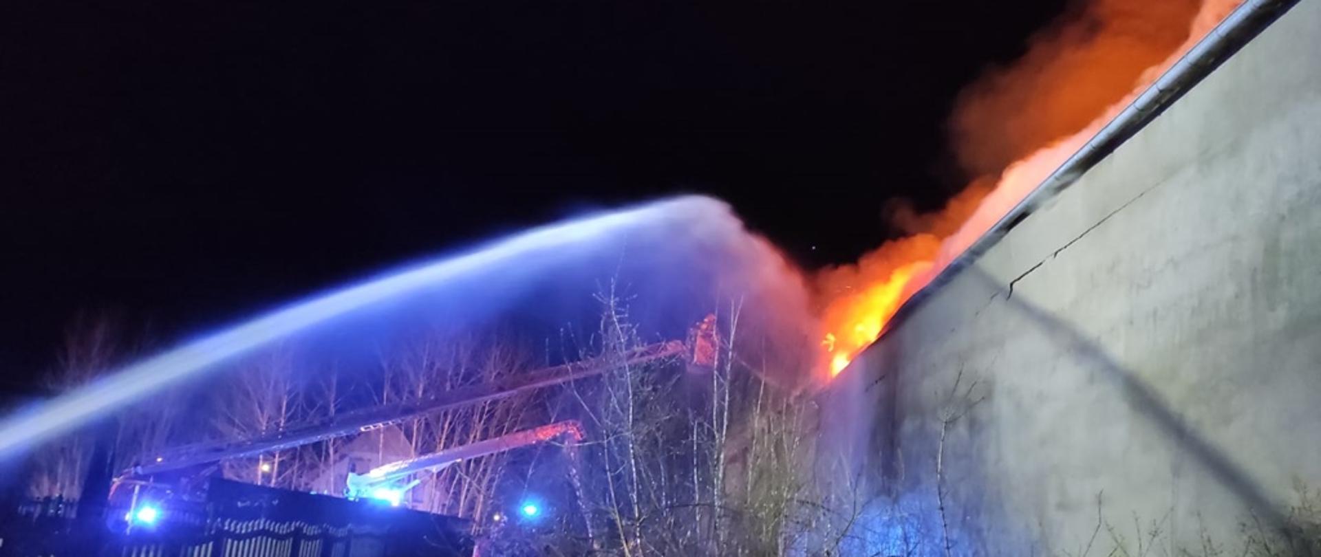po prawej stronie paląca się hala. po lewej dwa prądy wody podawane z drabin mechanicznych, jeden z działka wodnego.