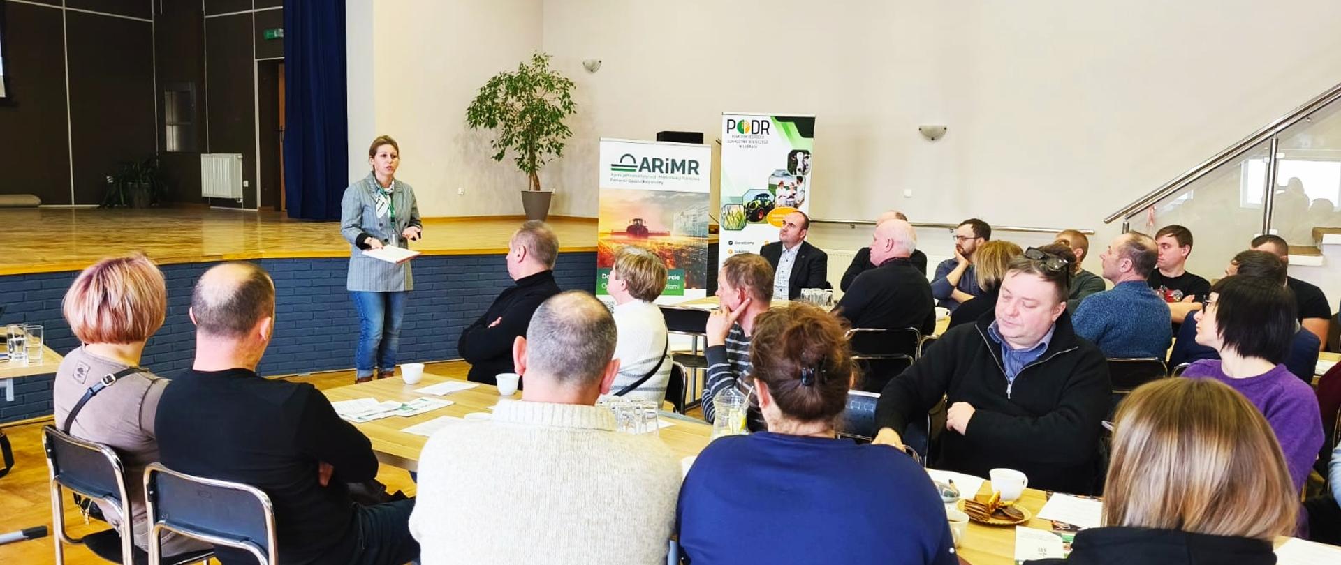 Sala ze sceną, przy stołach siedzą ludzie i słuchają wystąpienia kobiety, która stoi przodem do nich