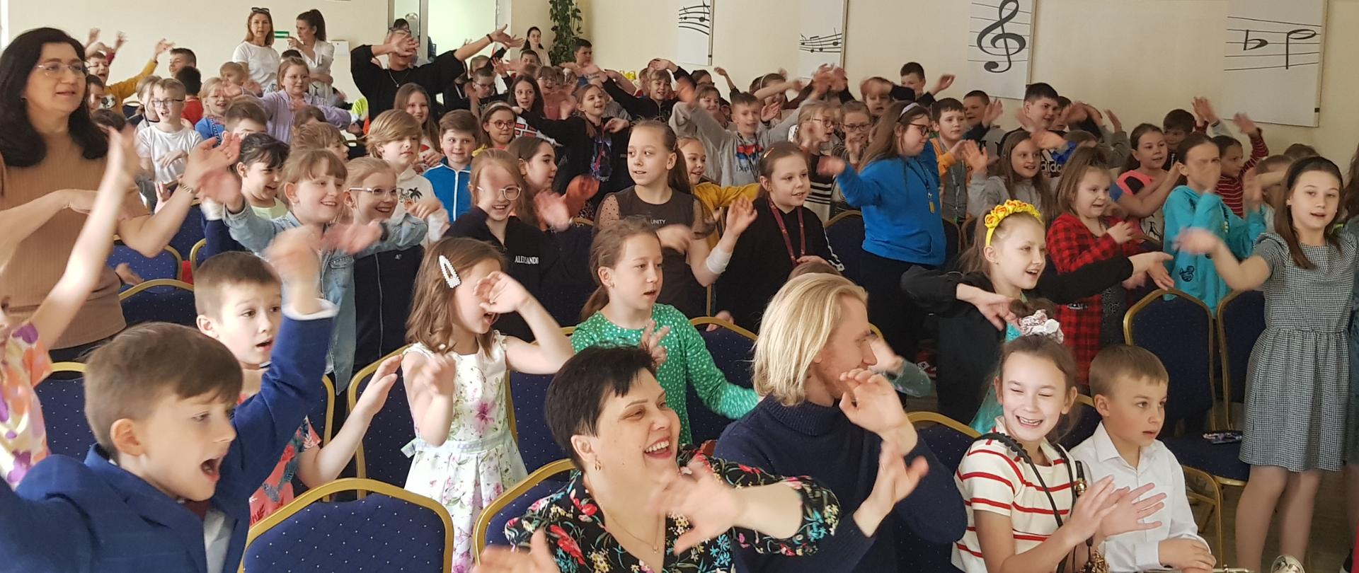 Zdjęcie przedstawia aulę PSM I st. w Grajewie i na niej dzieci tańczące, które uczestniczą w koncercie ,,Dźwięki Wiosny", dzieci są ubrane na kolorowo, mają uniesione dłonie. W tle zegar oraz grafiki z kluczem wiolinowym, pięciolinią i nutami w kolorach biało- czarnych. 