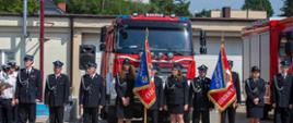 Poczty sztandarowe ochotniczych straży pożarnych stoją przed samochodami gaśniczymi podczas uroczystości.
