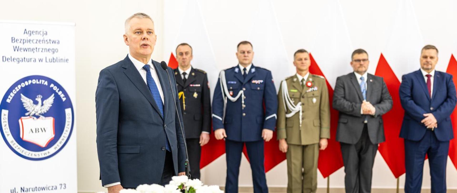 Minister Tomasz Siemoniak podczas przemówienia. Na pierwszym planie po lewej stronie biało czerwone kwiaty, nad nimi czarny mikrofon. W tle przedstawiciele służb RP. Za nimi biało-czerwone flagi. Po prawej stronie roll-up z napisem: Agencja Bezpieczeństwa Wewnętrznego Delegatura w Lublinie, na środku logo ABW, na dole napis: ul. Narutowicza 73 20-019 Lublin.