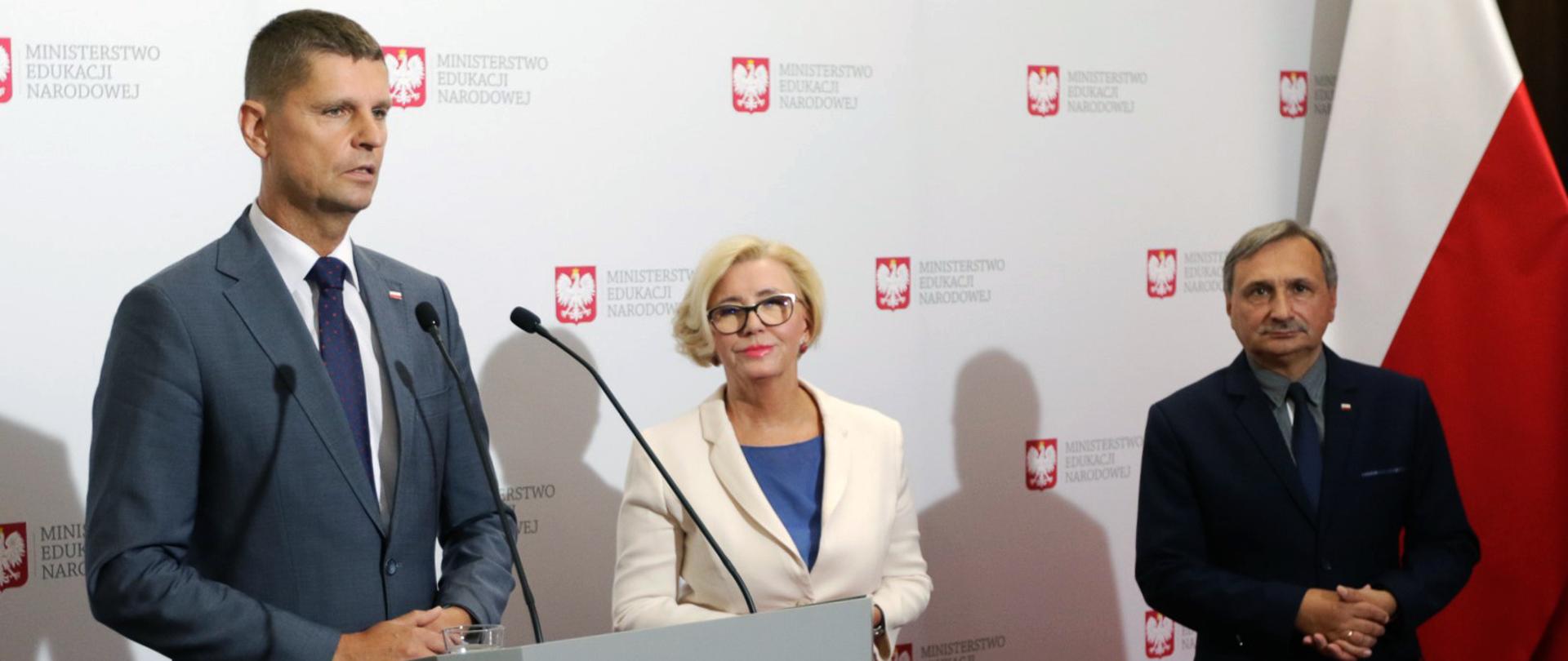 Konferencja prasowa Ministra Edukacji Narodowej Dariusza Piontkowskiego na temat bezpiecznego powrotu uczniów do szkół. Minister stoi za mównicą. Obok niego po prawej stronie stoją wiceministrowie. W tle biało-czerwona flaga. 