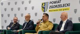 Na fotelach w kolorze zielonym siedzi 4 mężczyzn. Jeden jest funkcjonariuszem Państwowej Straży Pożarnej ubranym w mundur służbowy w kolorze piaskowym. W ręce trzyma mikrofon. Pozostali mężczyźni ubrani w garnitury. Mężczyźni prowadzą dyskusje. W tle ścianka informacyjna Powiatu Zgorzeleckiego.