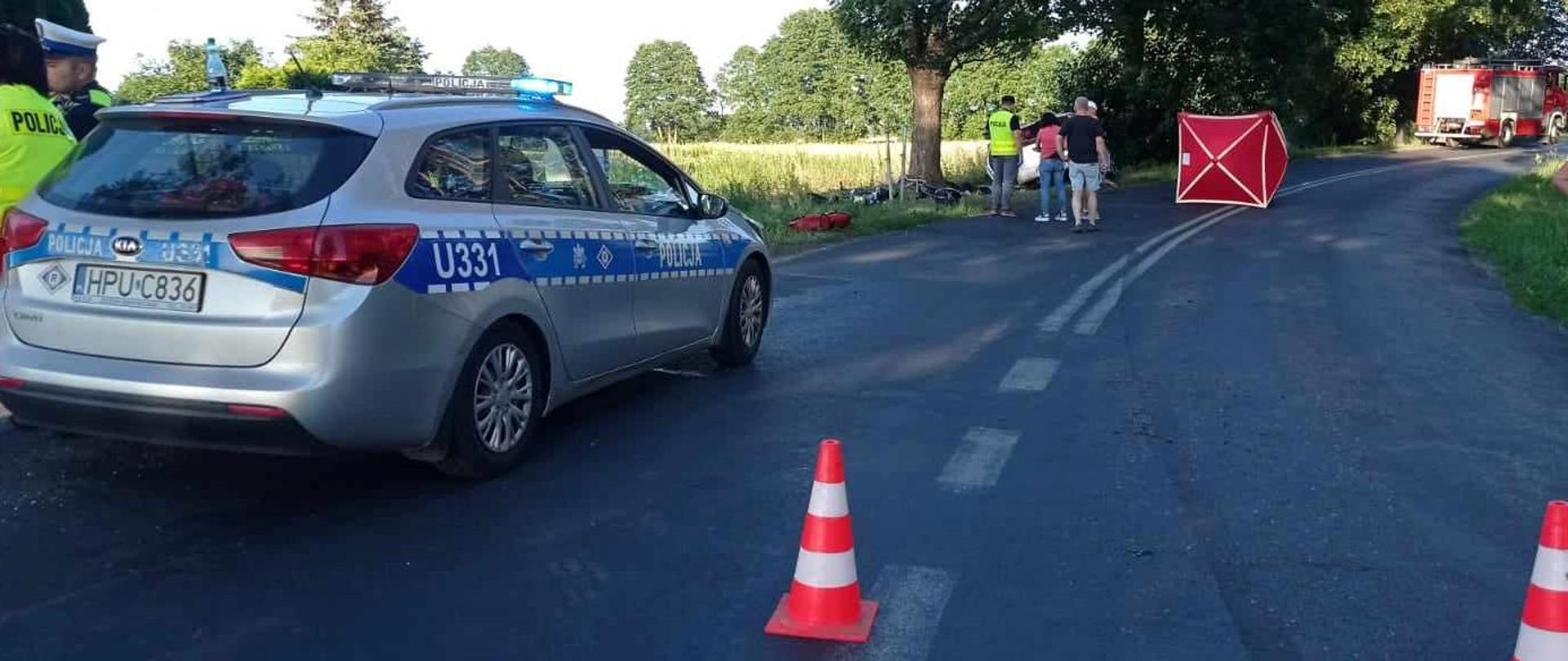 Zdjęcie przedstawia strażaków i policjantów podczas śmiertelnego wypadku motocyklisty.
W tle jednia, drzewa i pole.
