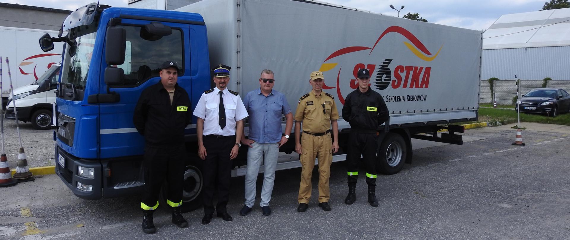 Na zdjęciu stoi pięciu mężczyzn. Od lewej pierwsza osoba to strażak ratownik Ochotniczej Straży Pożarnej w umundurowaniu koszarowym. Druga osoba od lewej w mundurze wyjściowym, w białej koszuli i ciemnych spodniach to starszy brygadier Paweł Malinowski - Zastępca Łódzkiego Komendanta Wojewódzkiego. Trzecia stojąca osoba to przedstawiciel ośrodka szkolenia kierowców w niebieskiej koszuli i jasnych spodniach. Czwartą stojącą osobą jest starszy brygadier Zbigniew Łyszkowicz - Zastępca Łódzkiego Komendanta Wojewódzkiego. Stoi on w umundurowaniu strażackim koloru piaskowego. Piąta stojąca osoba to drugi strażak ratownik Ochotniczej Straży Pożarnej w umundurowaniu koszarowym. Wszystkie pięć osób stoi na tle samochodu ciężarowego przeznaczonego do szkolenia kierowców.